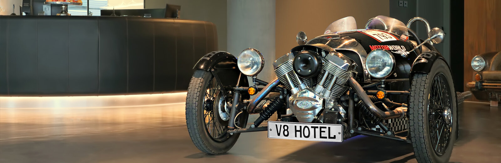 Ein klassisches, dreirädriges Fahrzeug steht im Eingangsbereich eines Hotels. Es hat eine schwarze Karosserie, glänzende Metallteile und einen Nummernschild mit der Aufschrift V8 HOTEL. Im Hintergrund ist der Empfangstresen zu sehen.