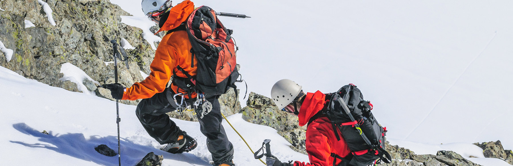 Zwei Bergsteiger in orangefarbener und roter Bekleidung klettern in verschneiter Umgebung. Sie verwenden Eispickel und Steigeisen, um steile Felsen und Schnee zu überwinden, während sie mit Rucksäcken ausgerüstet sind. Die Szene zeigt die Herausforderungen des Bergsteigens im Winter.